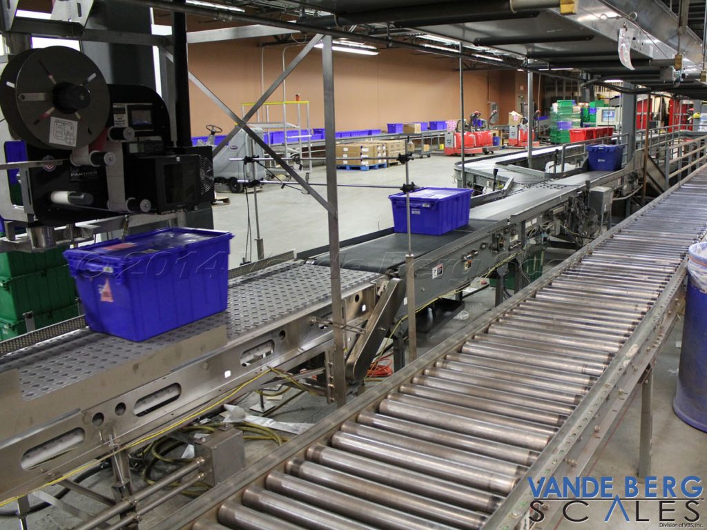 Plastic totes (of different heights) being labeled on the top. The system scans the height of the tote and adjusts the applicator arm accordingly.